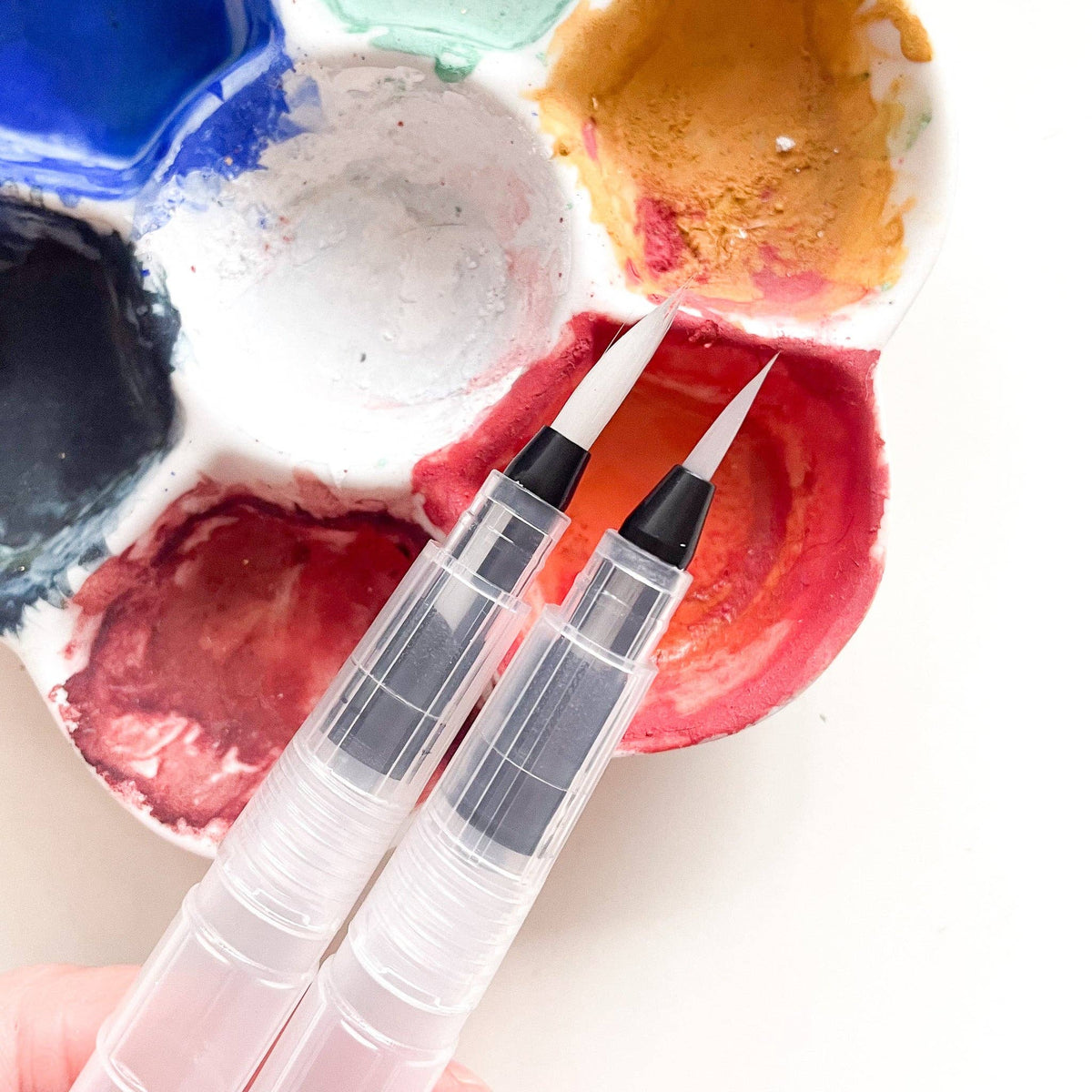 Two water brush tips resting in a palette of red, orange, blue and green paint