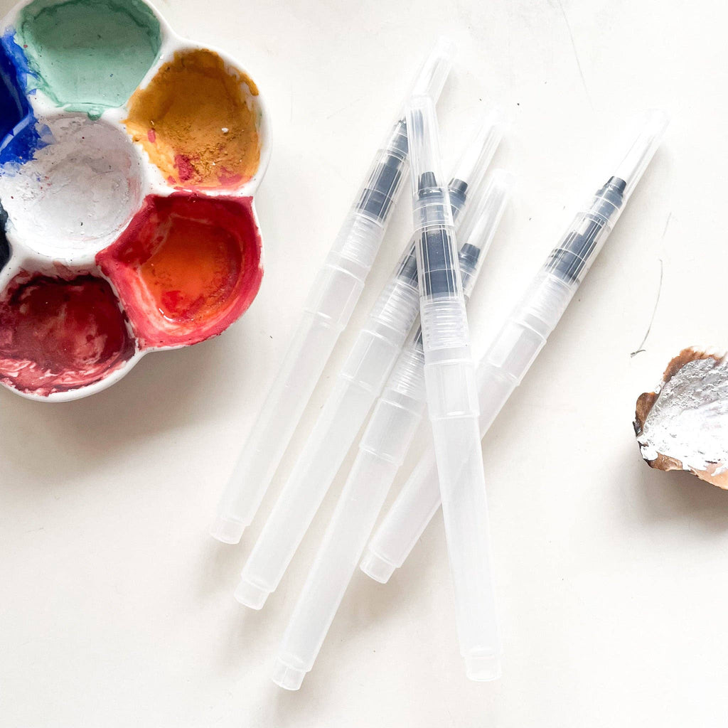 Several clear refillable water brushes beside a watercolor palette