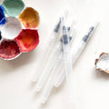 Several clear refillable water brushes beside a watercolor palette