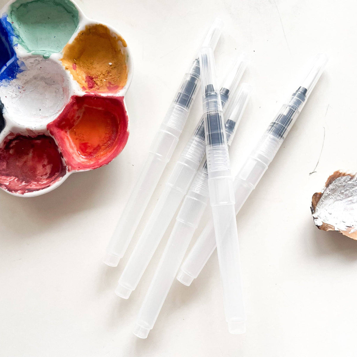 Several clear refillable water brushes beside a watercolor palette