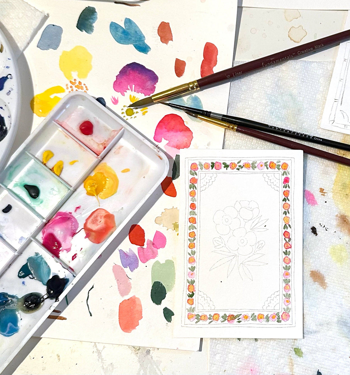 A floral border design in progress beside a paint palette and brushes