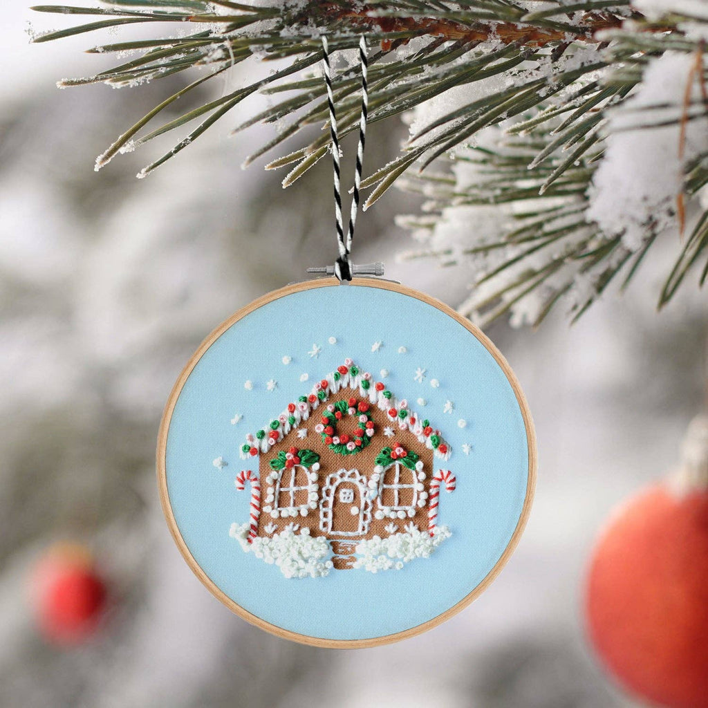 Finished embroidery hoop ornament of a gingerbread house on pale blue, hanging on a snowy pine