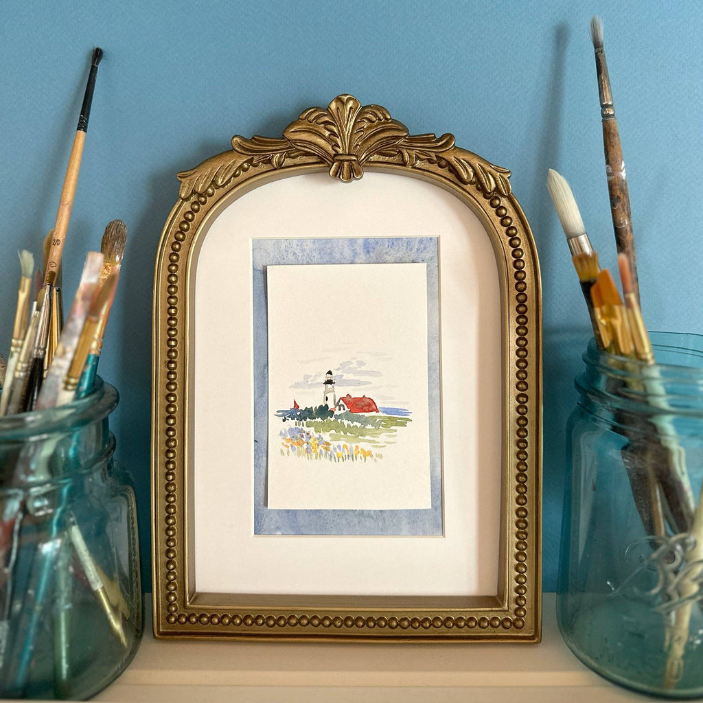 The finished lighthouse painting in a small gold arched frame, with brushes in jars