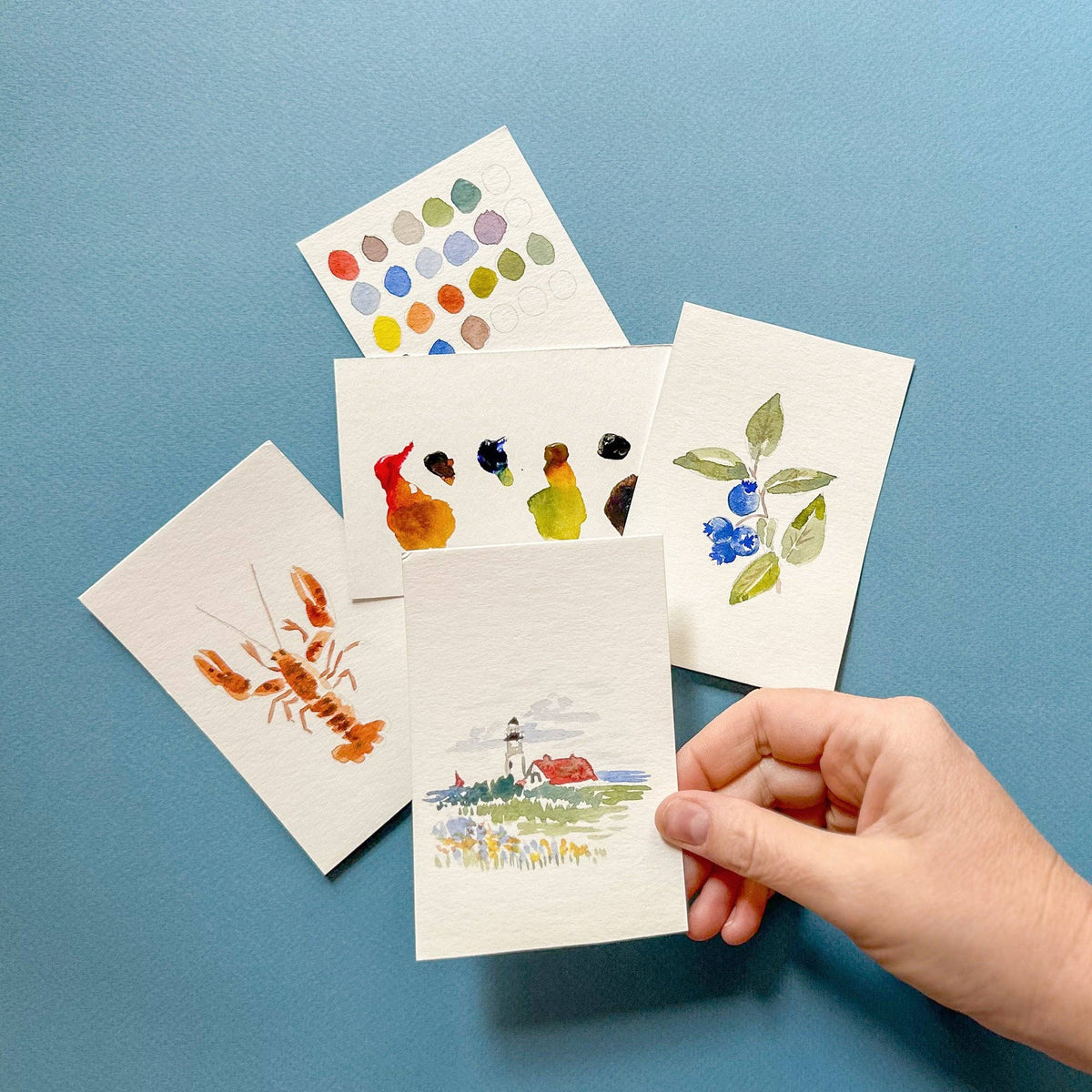 Hand holding the painted lighthouse card over the lobster, blueberry and paint cards