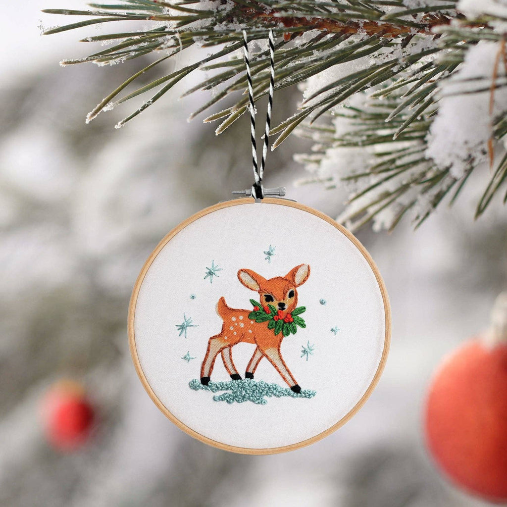 Finished embroidery hoop ornament of a fawn with a holly wreath, hanging on a snowy pine