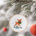 Finished embroidery hoop ornament of a fawn with a holly wreath, hanging on a snowy pine