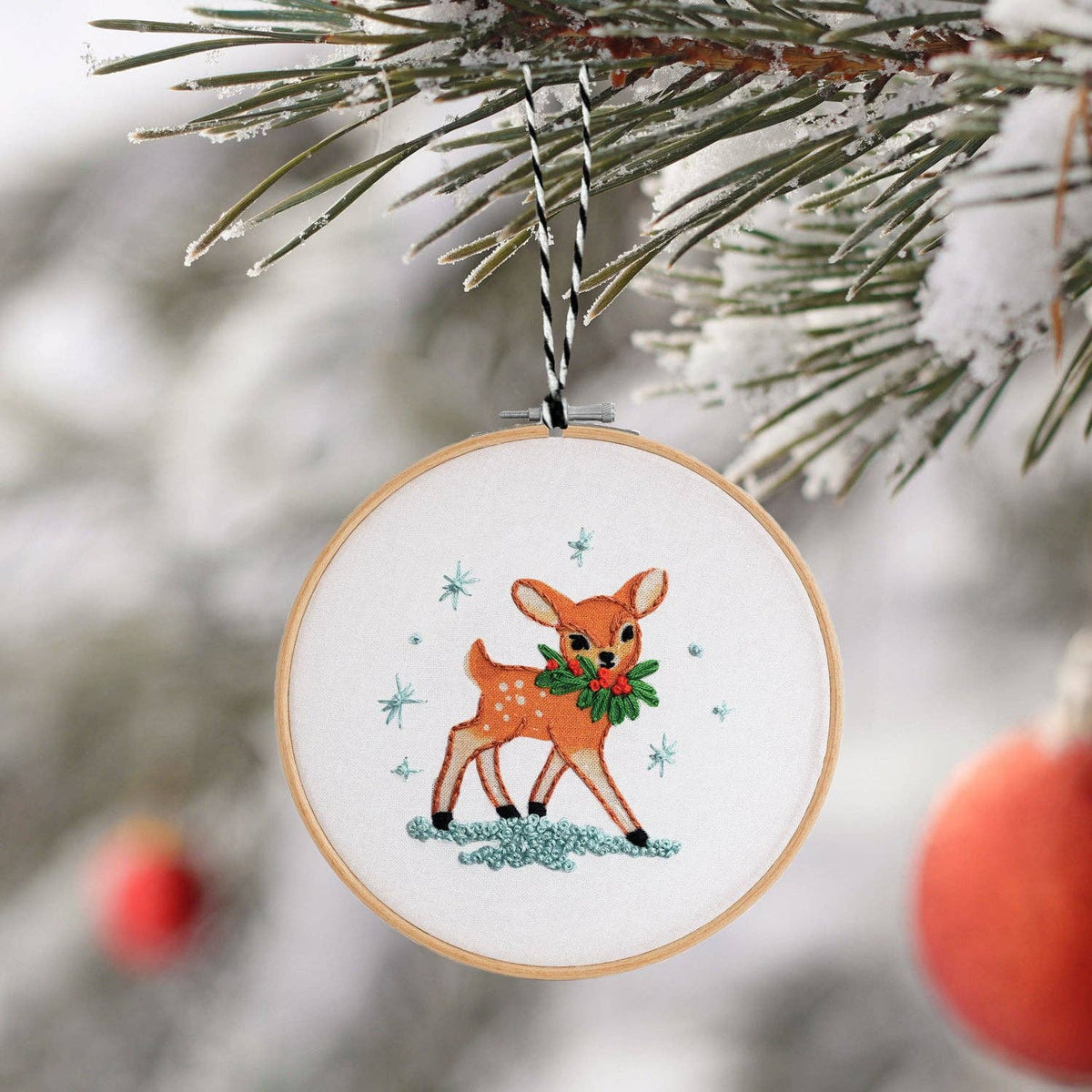 Finished embroidery hoop ornament of a fawn with a holly wreath, hanging on a snowy pine