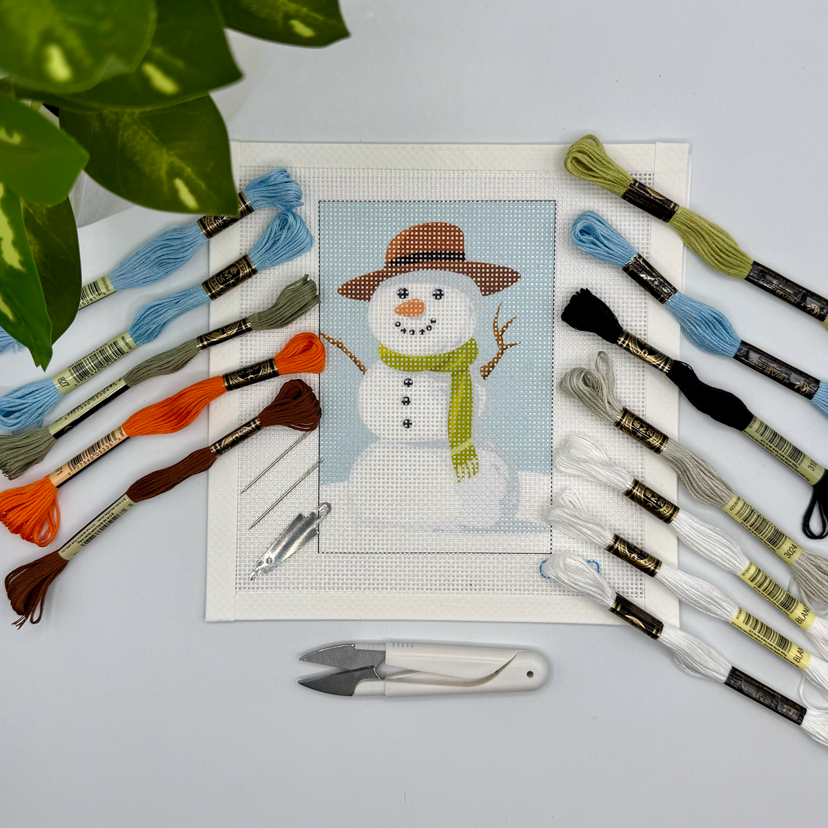 Snowman needlepoint kit: a painted canvas of a snowman in a brown hat and green scarf, with threads and tools