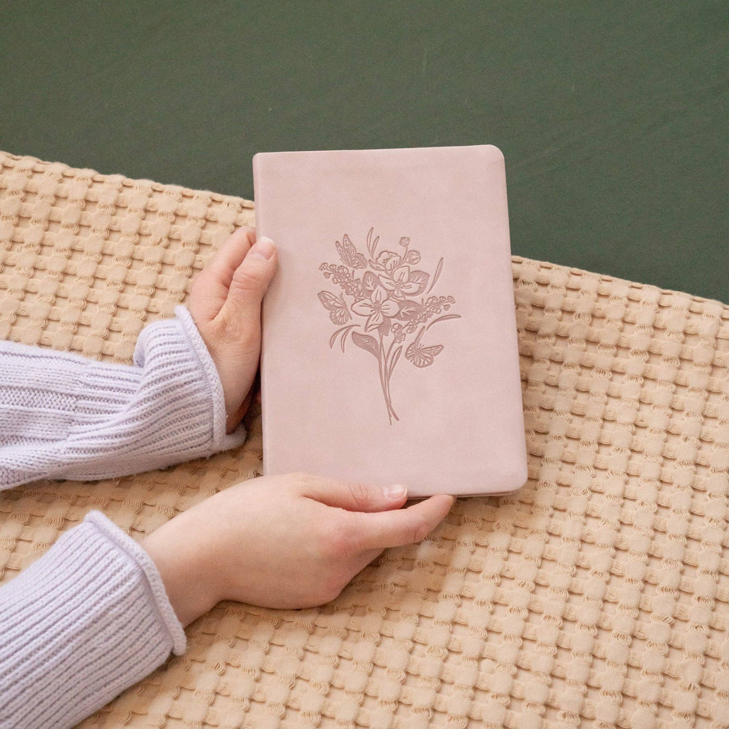 Hands holding the blush floral notebook on a woven blanket