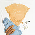 Mustard yellow tee with "be the light", Matthew 5:14, in white script, laid out with jeans