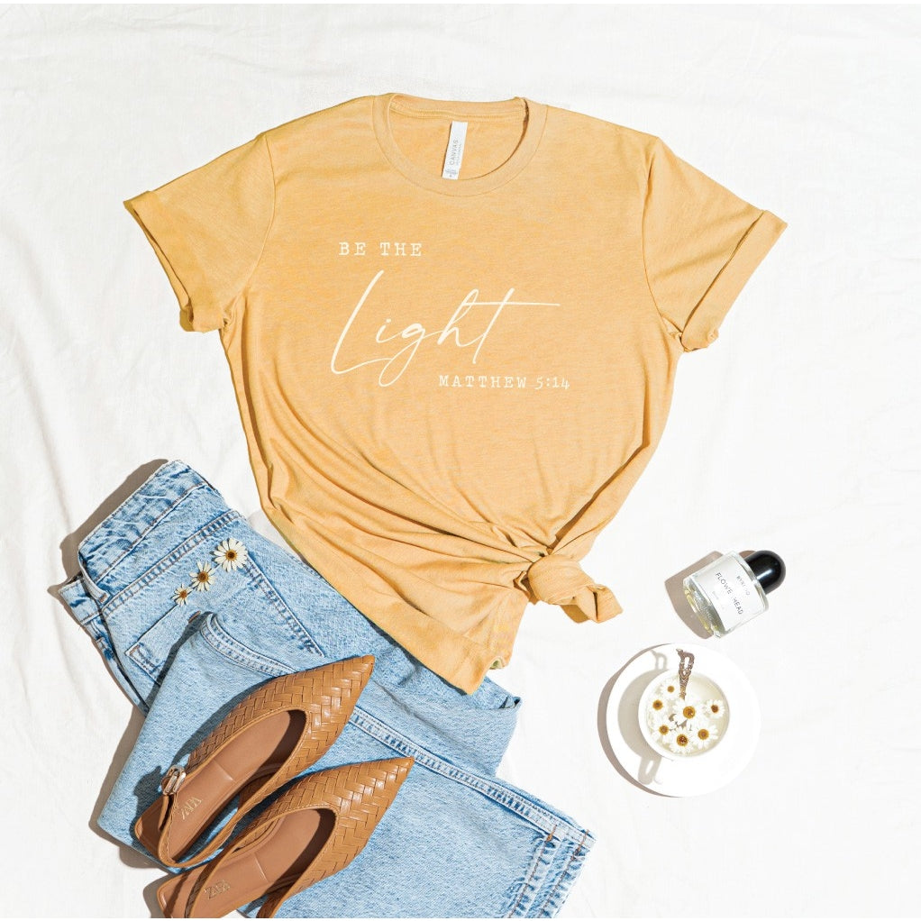 Mustard yellow tee with "be the light", Matthew 5:14, in white script, laid out with jeans