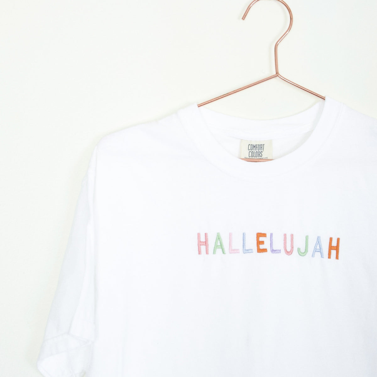 White tee with "HALLELUJAH" in multicolored pastel letters, on a hanger